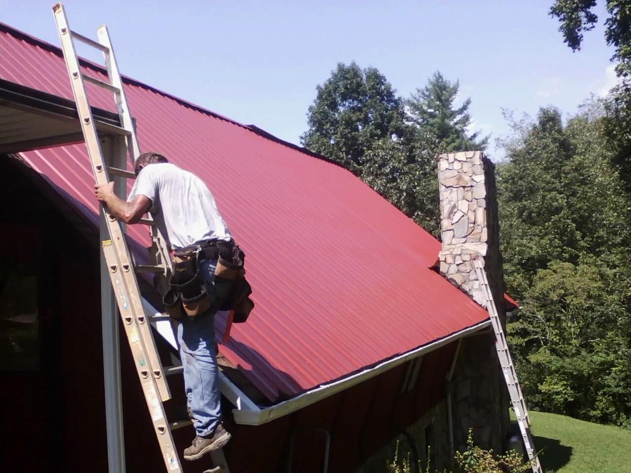 Roofer on a ladder beside a red metal roof and stone chimney — Soffit & Fascia Repair by Roofing Brothers in Lomita